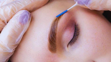 Um close-up da sobrancelha de uma pessoa sendo escovada com um pequeno aplicador por mãos usando luvas roxas; os olhos da pessoa estão fechados, capturando o cuidado preciso envolvido na laminação e micropigmentação da sobrancelha.