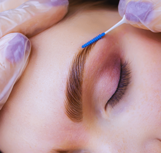 Um close-up da sobrancelha de uma pessoa sendo escovada com um pequeno aplicador por mãos usando luvas roxas; os olhos da pessoa estão fechados, capturando o cuidado preciso envolvido na laminação e micropigmentação da sobrancelha.