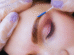 Um close-up da sobrancelha de uma pessoa sendo escovada com um pequeno aplicador por mãos usando luvas roxas; os olhos da pessoa estão fechados, capturando o cuidado preciso envolvido na laminação e micropigmentação da sobrancelha.