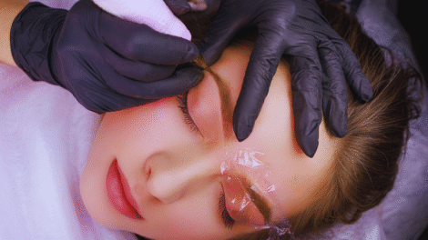 Uma pessoa com os olhos fechados recebe o tratamento de microblading na sobrancelha; uma mão enluvada garante a biossegurança ao segurar a ferramenta perto da sobrancelha, e um filme plástico protege a outra sobrancelha.