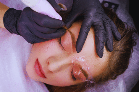 Uma pessoa com os olhos fechados recebe o tratamento de microblading na sobrancelha; uma mão enluvada garante a biossegurança ao segurar a ferramenta perto da sobrancelha, e um filme plástico protege a outra sobrancelha.