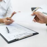 Duas pessoas se sentam em uma mesa. Uma entrega um envelope branco para a outra. Uma prancheta com um contrato e uma caneta está sobre a mesa entre elas. Somente suas mãos e braços estão visíveis.
