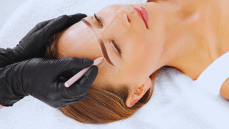 Uma pessoa com luvas pretas usa um lápis para delinear o formato das sobrancelhas no rosto de uma mulher, preparando-a para a micropigmentação de sobrancelhas enquanto ela se deita com os olhos fechados em uma toalha branca.