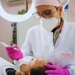 Uma micropigmentadora usando máscara e luvas se prepara para realizar um procedimento cosmético na área da sobrancelha de uma paciente em uma clínica.