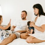 Uma jovem família está sentada em uma cama, sorrindo, com seu senso de parentesco evidente. O pai e a mãe, segurando um recém-nascido, observam a filha pequena tirar uma foto com o celular no quarto iluminado e aconchegante, adornado com roupas de cama brancas e um travesseiro floral.