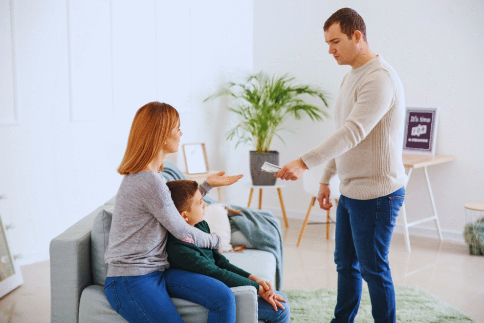 Um homem entrega dinheiro a uma mulher sentada que está com uma criança em uma sala de estar, ilustrando o pagamento da pensão alimentícia retroativa.