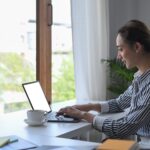 Uma mulher com uma camisa listrada senta-se em uma mesa perto de uma janela, trabalhando em um laptop. Há uma xícara de café, artigos de papelaria e uma planta sobre a mesa branca, com luz natural enchendo o ambiente.