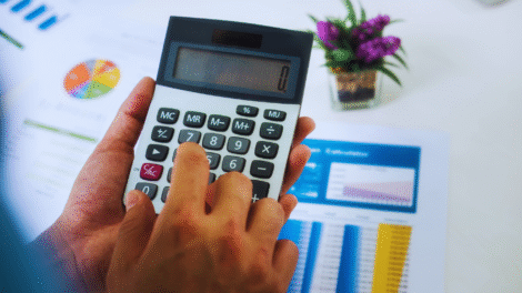 Uma pessoa usa uma calculadora, com gráficos financeiros impressos e um pequeno vaso de planta visível em uma mesa branca - talvez analisando qual é o melhor investimento hoje.