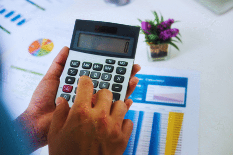 Uma pessoa usa uma calculadora, com gráficos financeiros impressos e um pequeno vaso de planta visível em uma mesa branca - talvez analisando qual é o melhor investimento hoje.