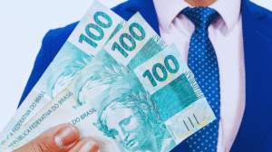 Uma pessoa de terno azul e gravata segura três notas de 100 reais, considerando onde investir 1.000 reais com sabedoria.