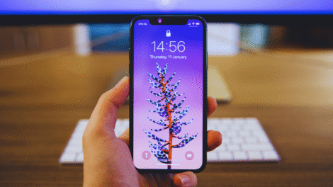 Uma pessoa segura um smartphone que exibe uma tela de bloqueio roxa com o desenho de uma planta; um teclado e um monitor são visíveis no fundo desfocado, sugerindo uma configuração de comparação entre o S23 Ultra e o iPhone 14 Pro Max.