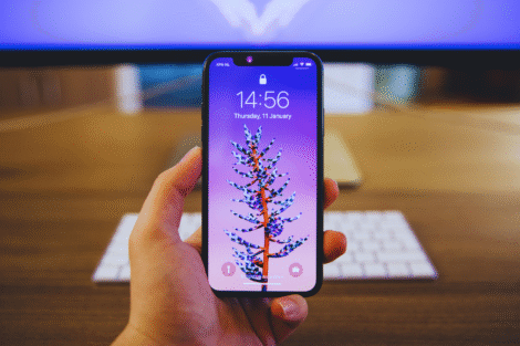 Uma pessoa segura um smartphone que exibe uma tela de bloqueio roxa com o desenho de uma planta; um teclado e um monitor são visíveis no fundo desfocado, sugerindo uma configuração de comparação entre o S23 Ultra e o iPhone 14 Pro Max.
