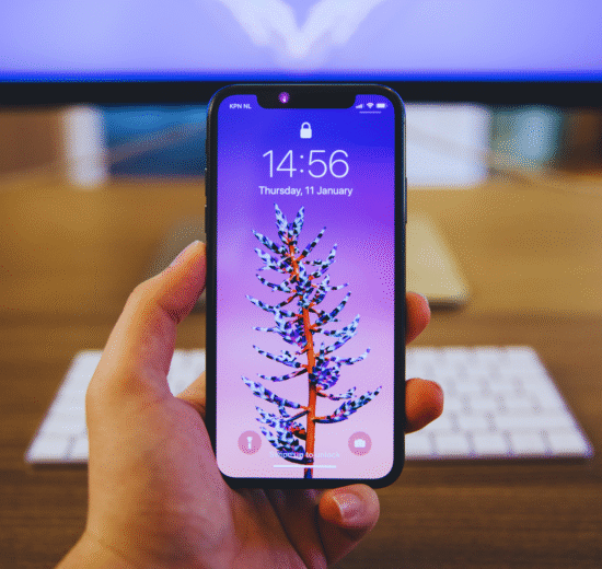 Uma pessoa segura um smartphone que exibe uma tela de bloqueio roxa com o desenho de uma planta; um teclado e um monitor são visíveis no fundo desfocado, sugerindo uma configuração de comparação entre o S23 Ultra e o iPhone 14 Pro Max.