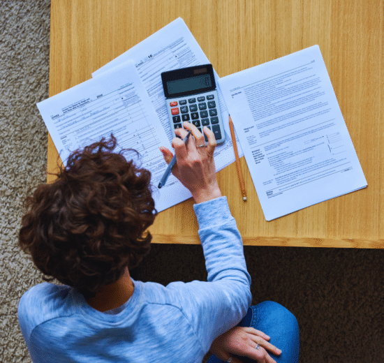 Pessoa sentada em uma mesa usando uma calculadora com vários documentos e formulários de imposto de renda 2023 espalhados à sua frente, procurando por dicas sobre como pagar menos imposto de renda.