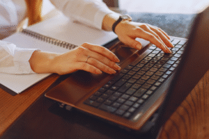 Pessoa digitando em um laptop com Windows 11 e um notebook aberto próximo a uma mesa de madeira. Somente as mãos e parte dos braços estão visíveis.