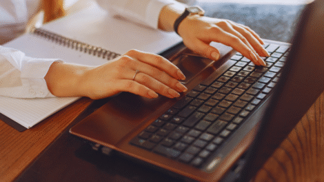 Pessoa digitando em um laptop com Windows 11 e um notebook aberto próximo a uma mesa de madeira. Somente as mãos e parte dos braços estão visíveis.