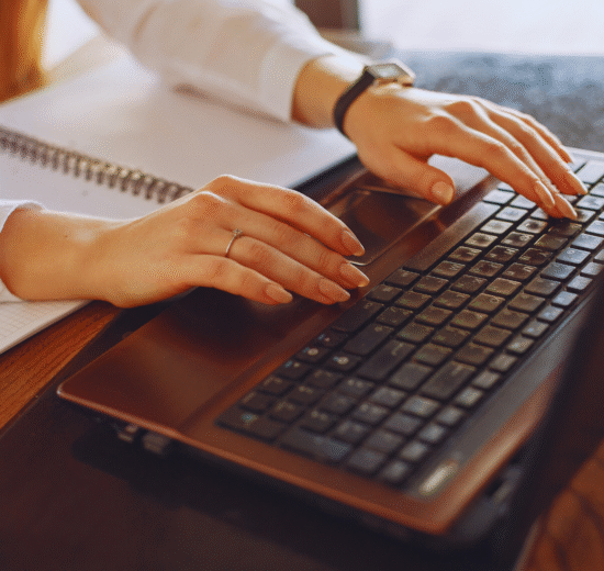 Pessoa digitando em um laptop com Windows 11 e um notebook aberto próximo a uma mesa de madeira. Somente as mãos e parte dos braços estão visíveis.