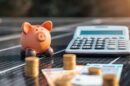 Um cofrinho, uma calculadora, pilhas de moedas e notas são colocados em um painel solar, simbolizando a economia financeira e o investimento em energia renovável.