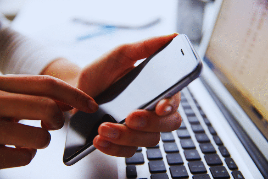 Uma pessoa usando um smartphone em uma mão enquanto está sentada em frente a um teclado de laptop, procurando informações sobre o Atestado 15 dias INSS.