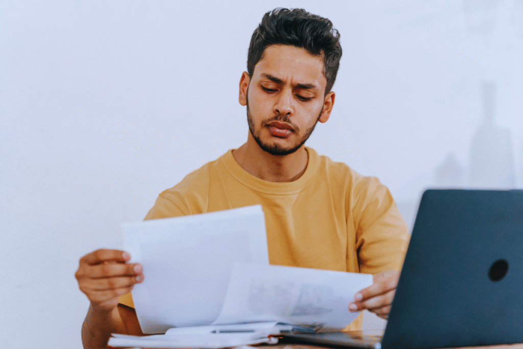 Um homem de camisa amarela está sentado em uma mesa, olhando para documentos CAT com uma expressão concentrada, ao lado de um laptop aberto.