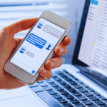 Uma pessoa segura um smartphone exibindo um chatbot do WhatsApp, ao lado de um laptop aberto mostrando uma caixa de entrada de e-mail, ilustrando como implementar soluções de mensagens entre plataformas.
