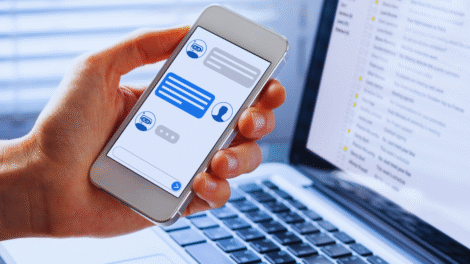 Uma pessoa segura um smartphone exibindo um chatbot do WhatsApp, ao lado de um laptop aberto mostrando uma caixa de entrada de e-mail, ilustrando como implementar soluções de mensagens entre plataformas.