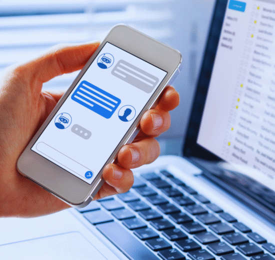 Uma pessoa segura um smartphone exibindo um chatbot do WhatsApp, ao lado de um laptop aberto mostrando uma caixa de entrada de e-mail, ilustrando como implementar soluções de mensagens entre plataformas.