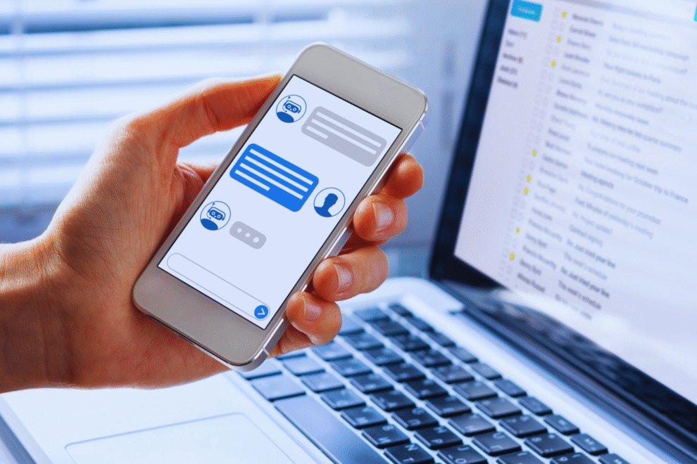 Uma pessoa segura um smartphone exibindo um chatbot do WhatsApp, ao lado de um laptop aberto mostrando uma caixa de entrada de e-mail, ilustrando como implementar soluções de mensagens entre plataformas.