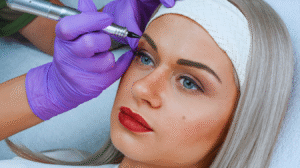 Uma pessoa com cabelos loiros está recebendo um procedimento cosmético de tatuagem de sobrancelha de um profissional usando luvas roxas, destacando a importância de seguir os cuidados pós micropigmentação para obter melhores resultados.