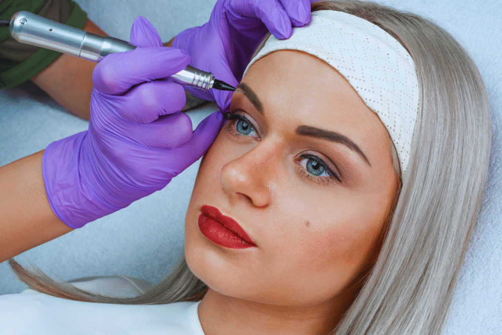 Uma pessoa com cabelos loiros está recebendo um procedimento cosmético de tatuagem de sobrancelha de um profissional usando luvas roxas, destacando a importância de seguir os cuidados pós micropigmentação para obter melhores resultados.
