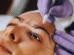 Uma pessoa recebe marcações nas sobrancelhas com uma caneta como parte de um procedimento de nanoblading sobrancelha; mãos usando luvas roxas seguram seu rosto com firmeza.